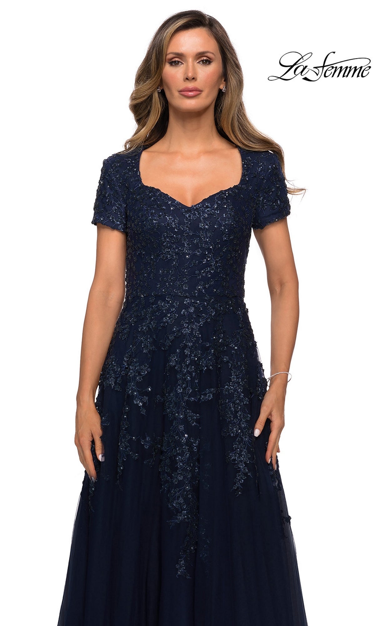 A woman wears a navy blue Long Formal Dress: La Femme 28037 featuring a sweetheart neckline, short sleeves, and intricate beaded appliques on the bodice over an A-line tulle skirt.